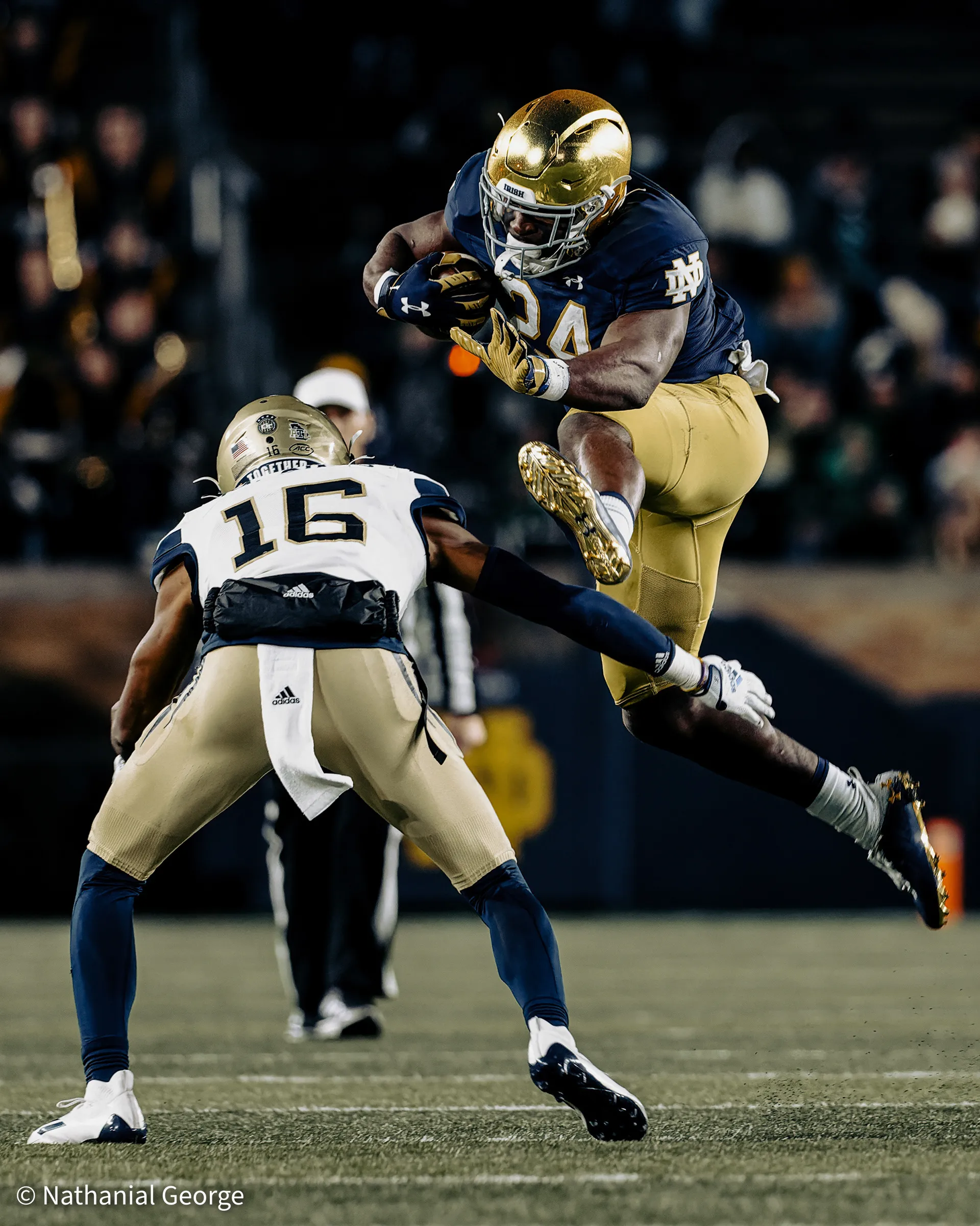 A Notre Dame ball carrier in navy and a gold helmet, number 24, leaps with the ball tucked as a defender in white, number 16, sets his feet to meet him.