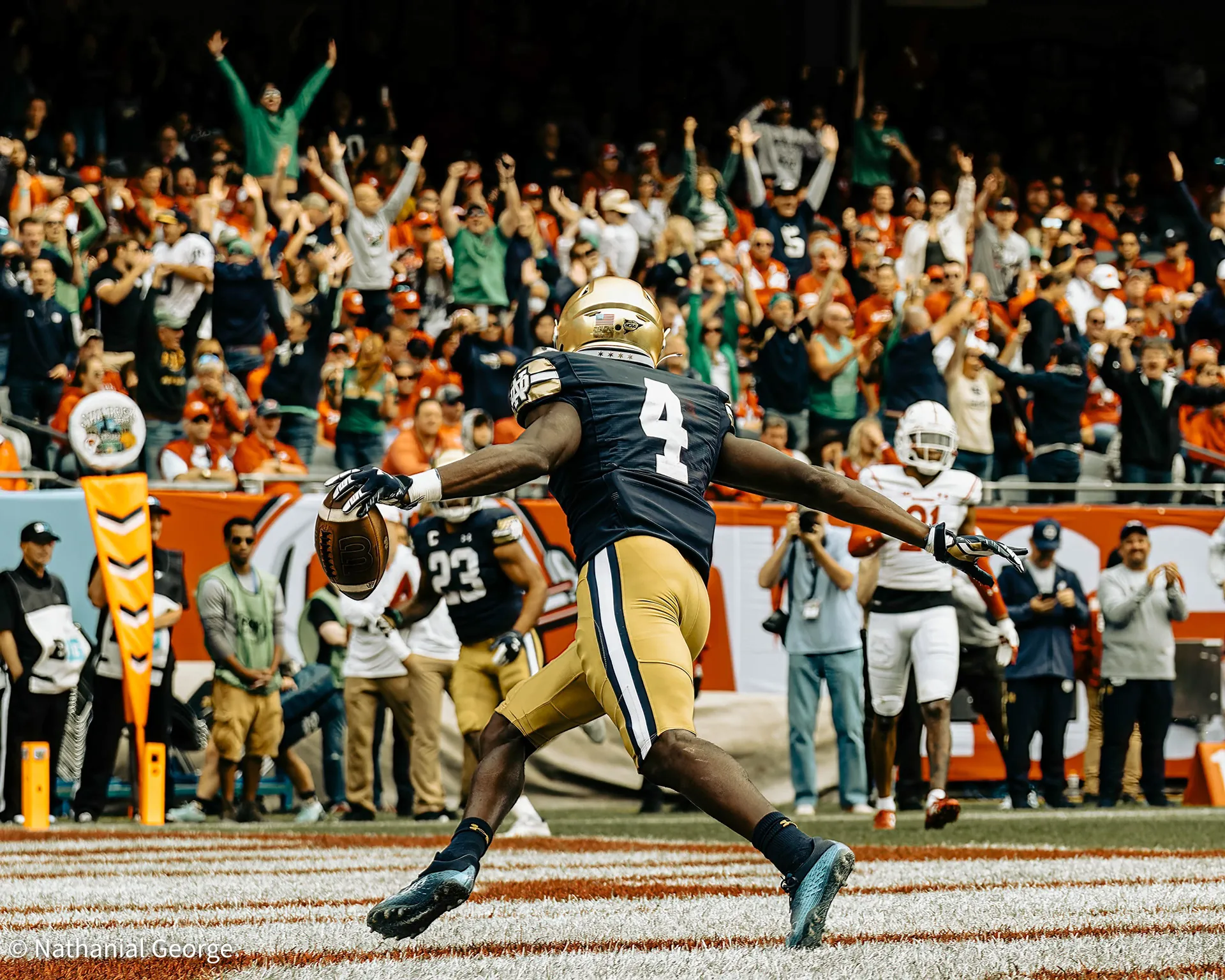 A Notre Dame player in navy, number 4, spreads his arms with the ball in one hand as he crosses the striped end zone, fans behind him throwing their arms up.