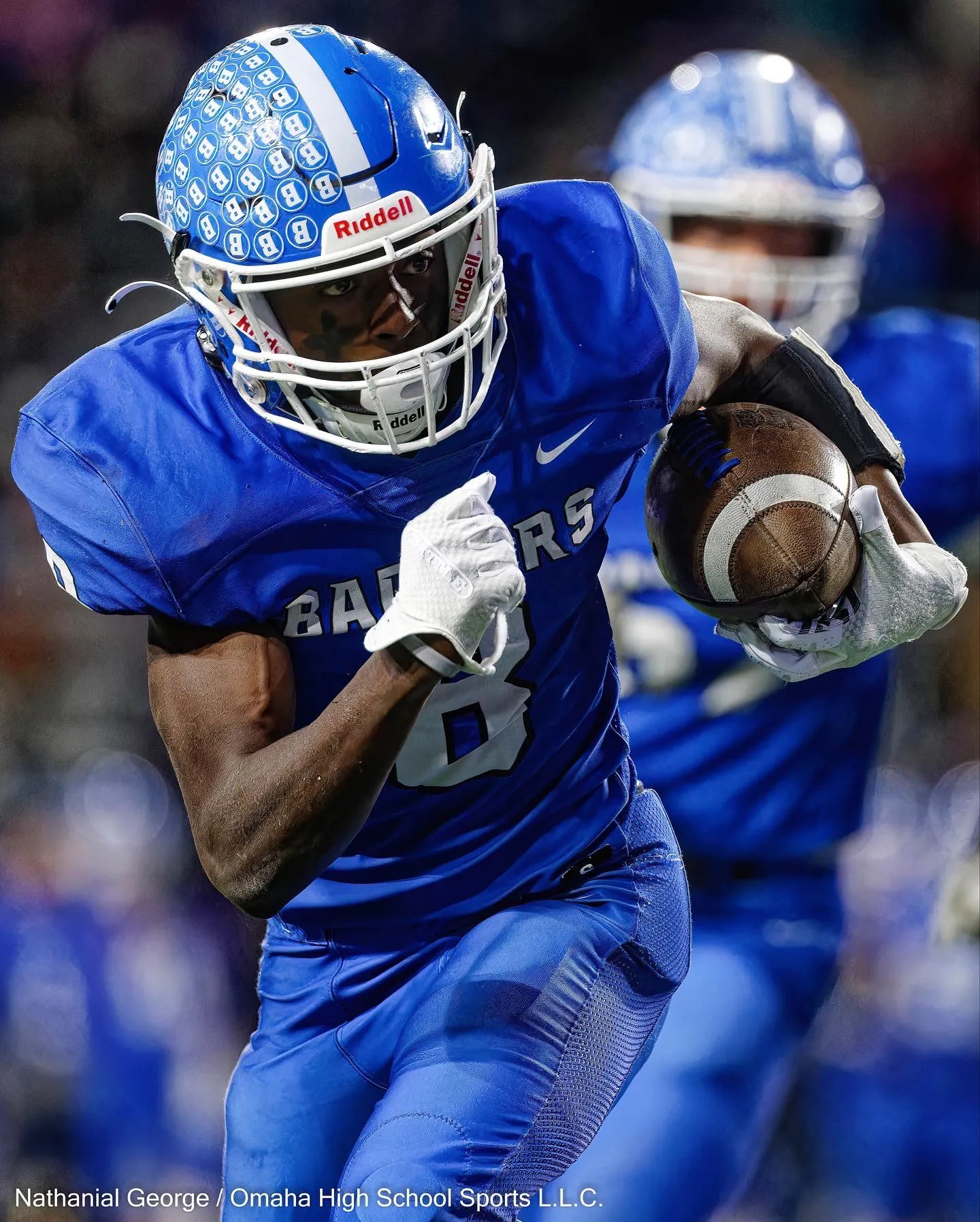 A ball carrier in royal blue, number 8, with small B marks covering his helmet, runs with the ball tucked against his side as a teammate follows out of focus.
