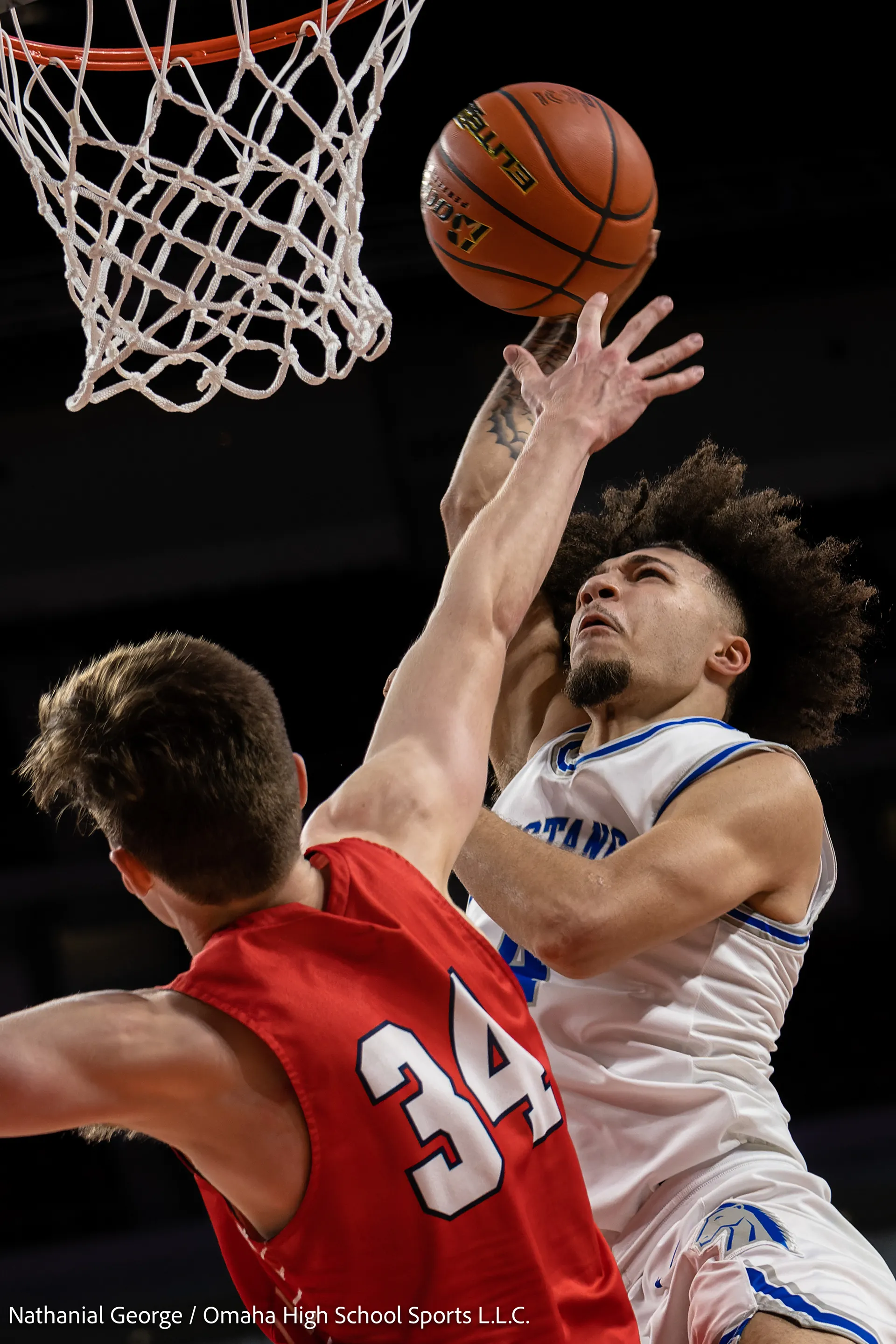 A player in white and blue rises to the basket with the ball above the rim while a defender in red, number 34, stretches an arm up to contest the shot.