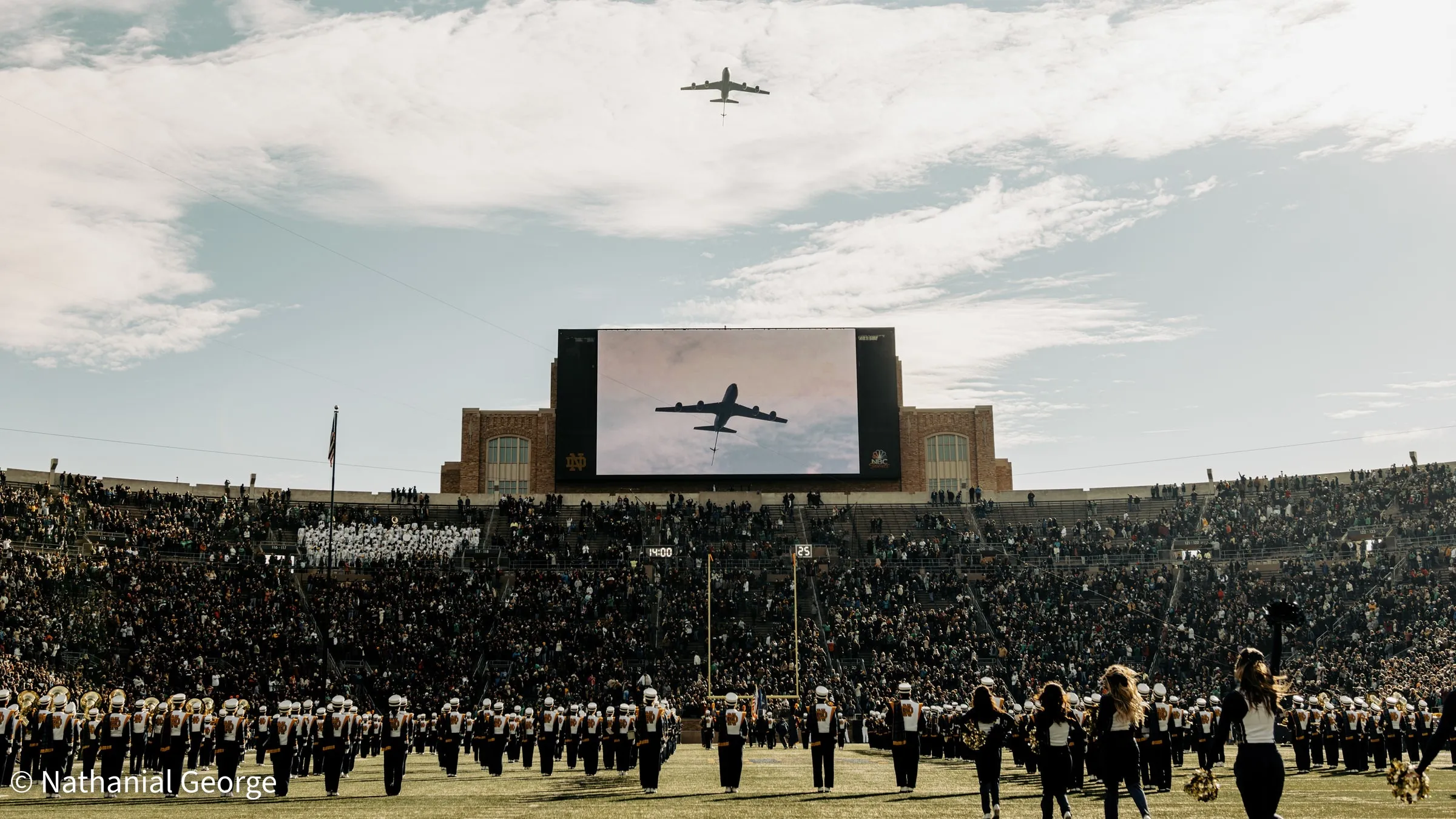 A large jet flies over a packed stadium while the video board below shows the same plane, a marching band stands in formation on the field and cheerleaders face the crowd in the foreground.