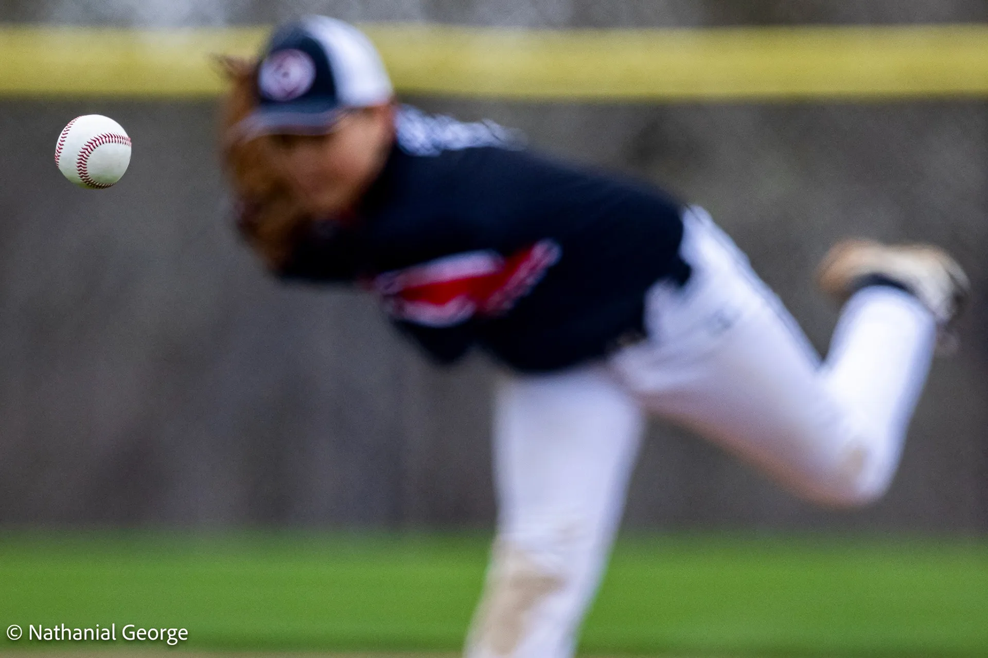 A baseball hangs in sharp focus at the left of the frame while the pitcher who threw it, in navy and white, is a soft blur in his follow-through.