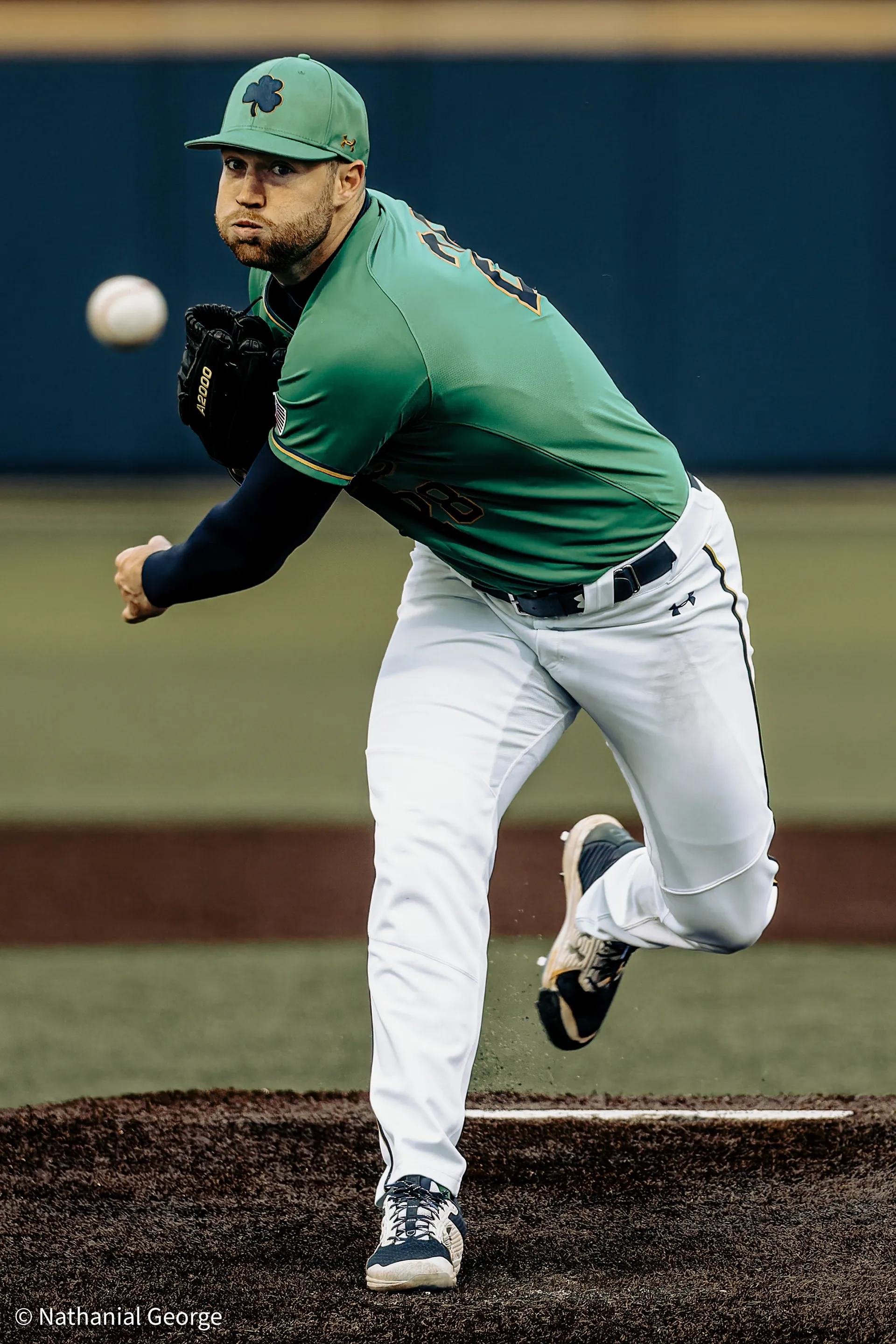 A pitcher in a green jersey and a green cap with a shamrock drives off the mound, the ball just out of his hand at the left of the frame.