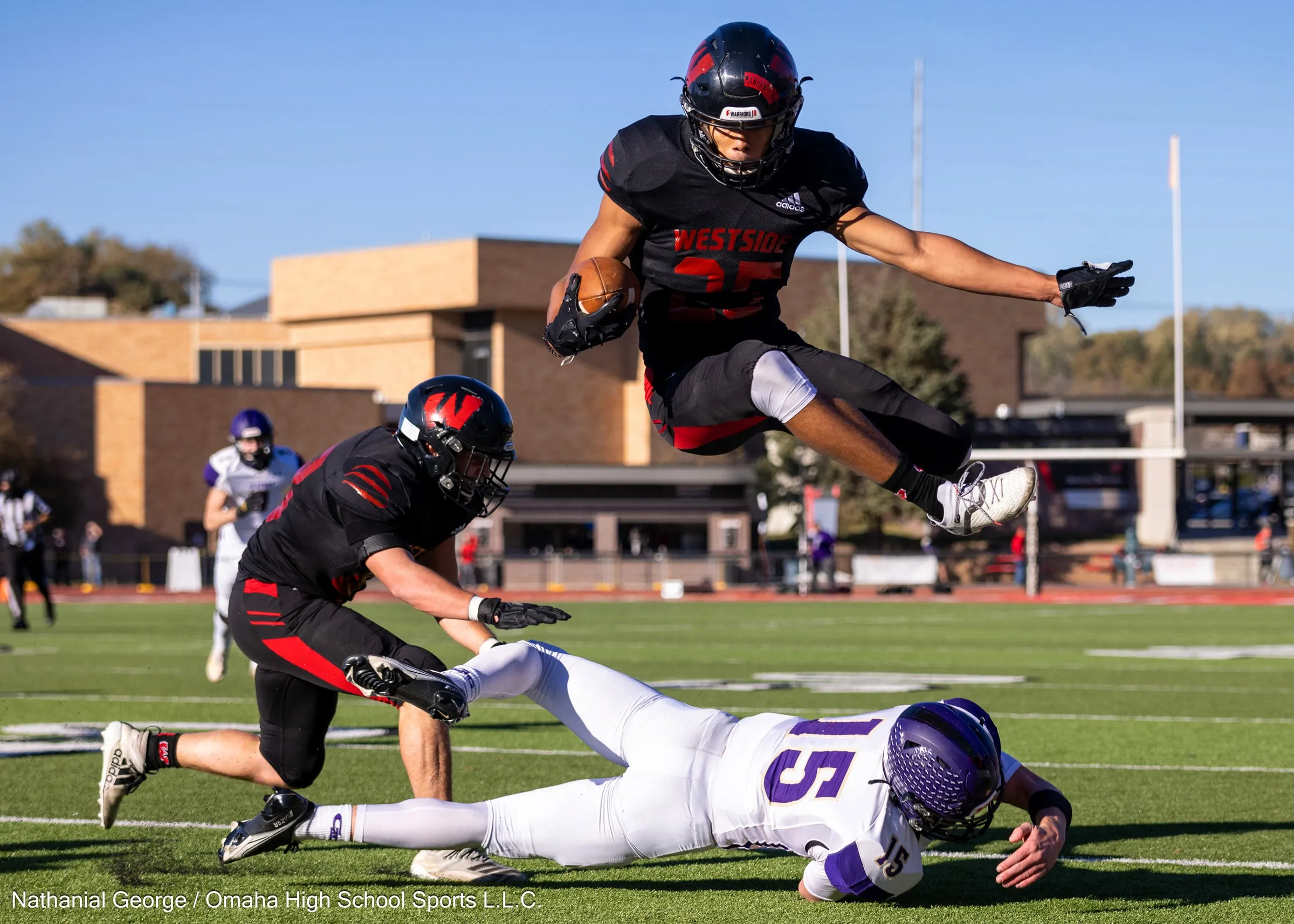 A Westside ball carrier in black and red, number 25, leaps over a defender in white and purple diving at his feet, a teammate beside him, on a sunny afternoon with a brick building behind.