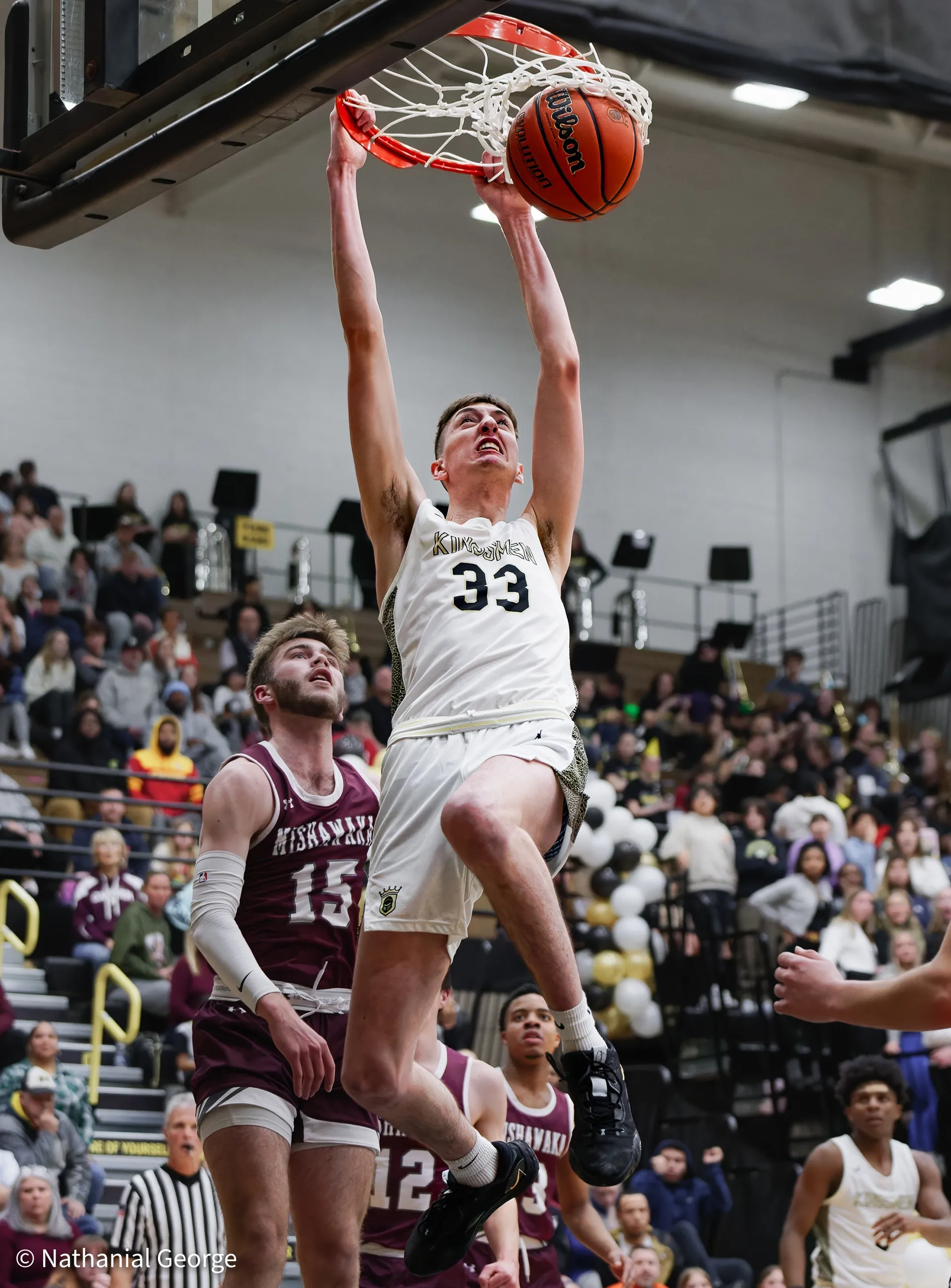 A player in white, number 33, hangs from the rim after a dunk while a defender in maroon, number 15, looks up at him, a full gym behind them.