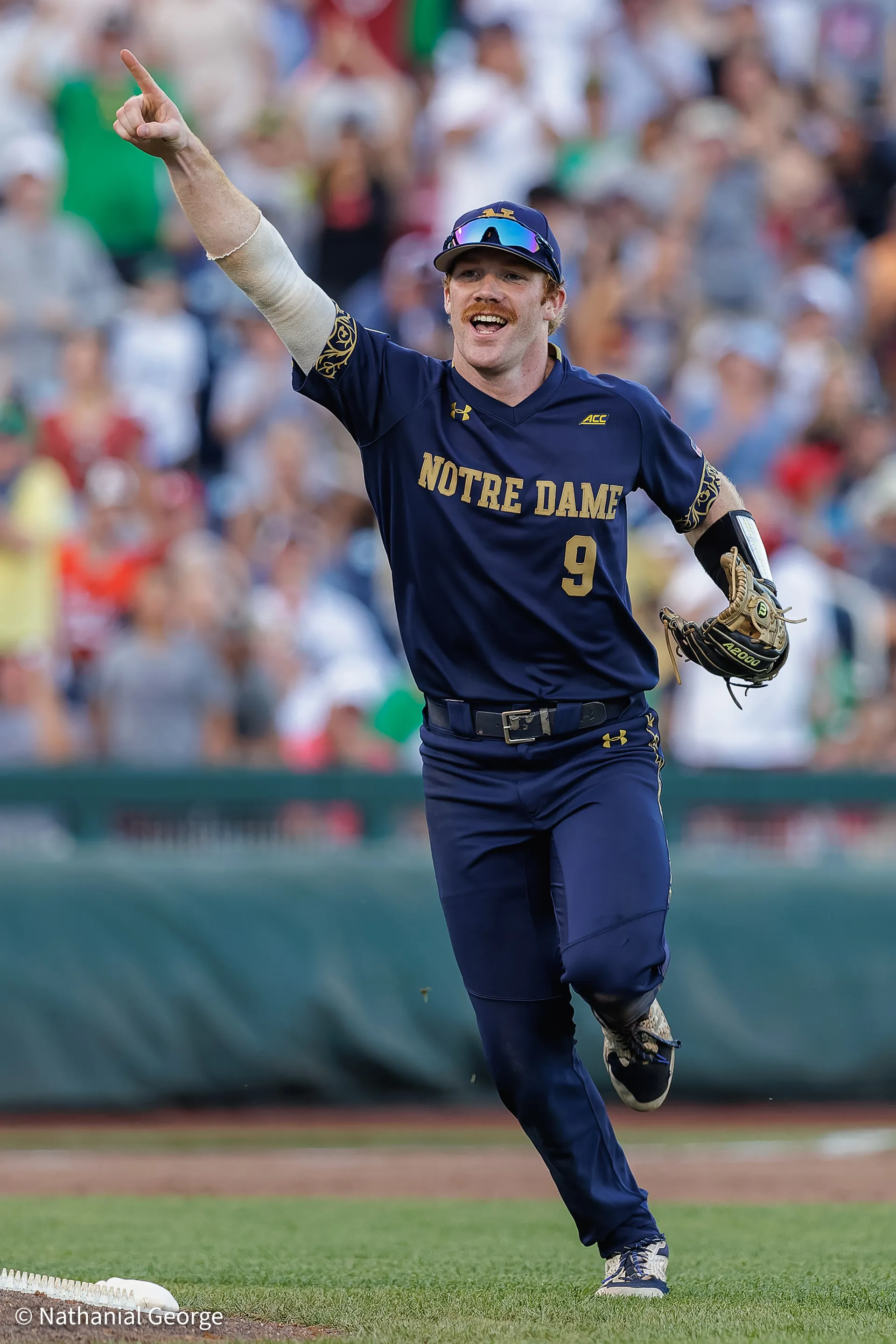 A Notre Dame infielder in navy, number 9, points skyward and shouts as he runs off the field, his glove on one hand and a packed crowd behind him.