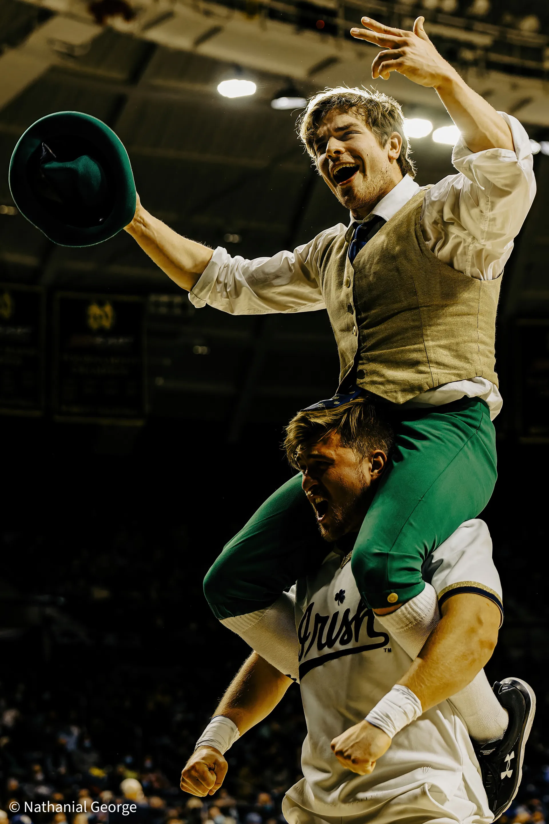 The Notre Dame leprechaun, his green hat held out in one hand, rides on the shoulders of a cheerleader in a white Irish jersey, both shouting under the arena lights.