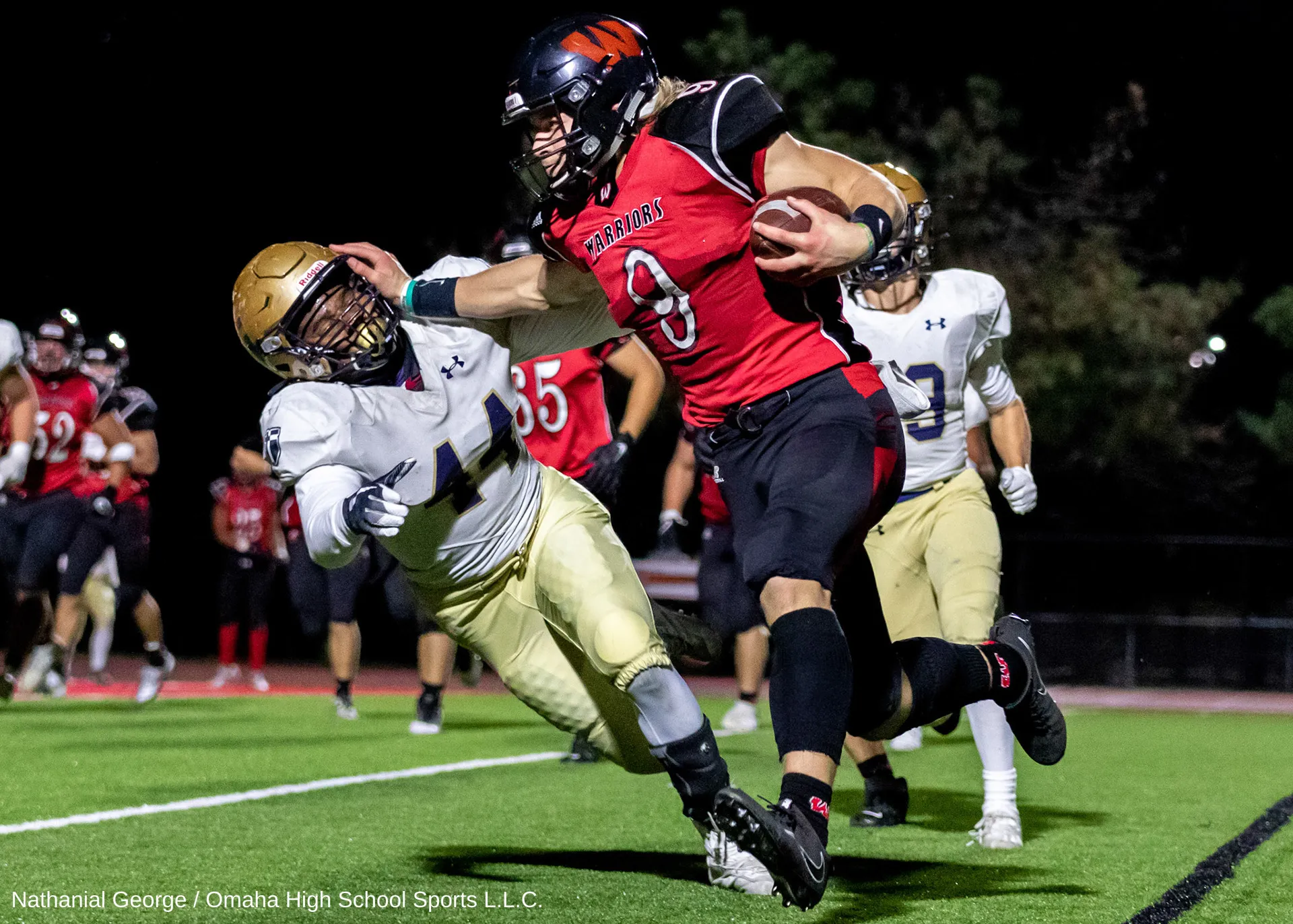 A Warriors ball carrier in red, number 8, pushes away a defender in white and gold with a straight arm as he runs along the edge of the field at night.