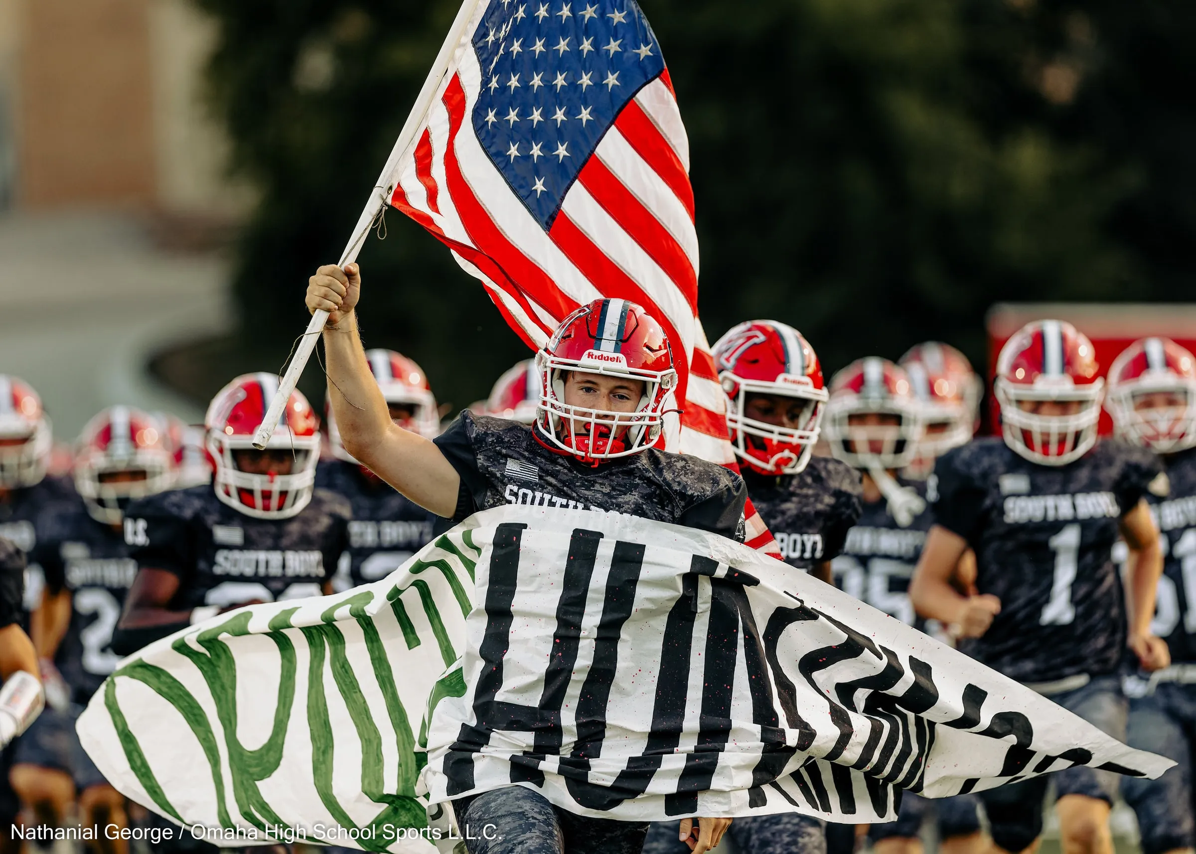 A player carries an American flag as his team, in dark camouflage-pattern jerseys and red helmets, runs onto the field behind a paper banner painted in green and black.
