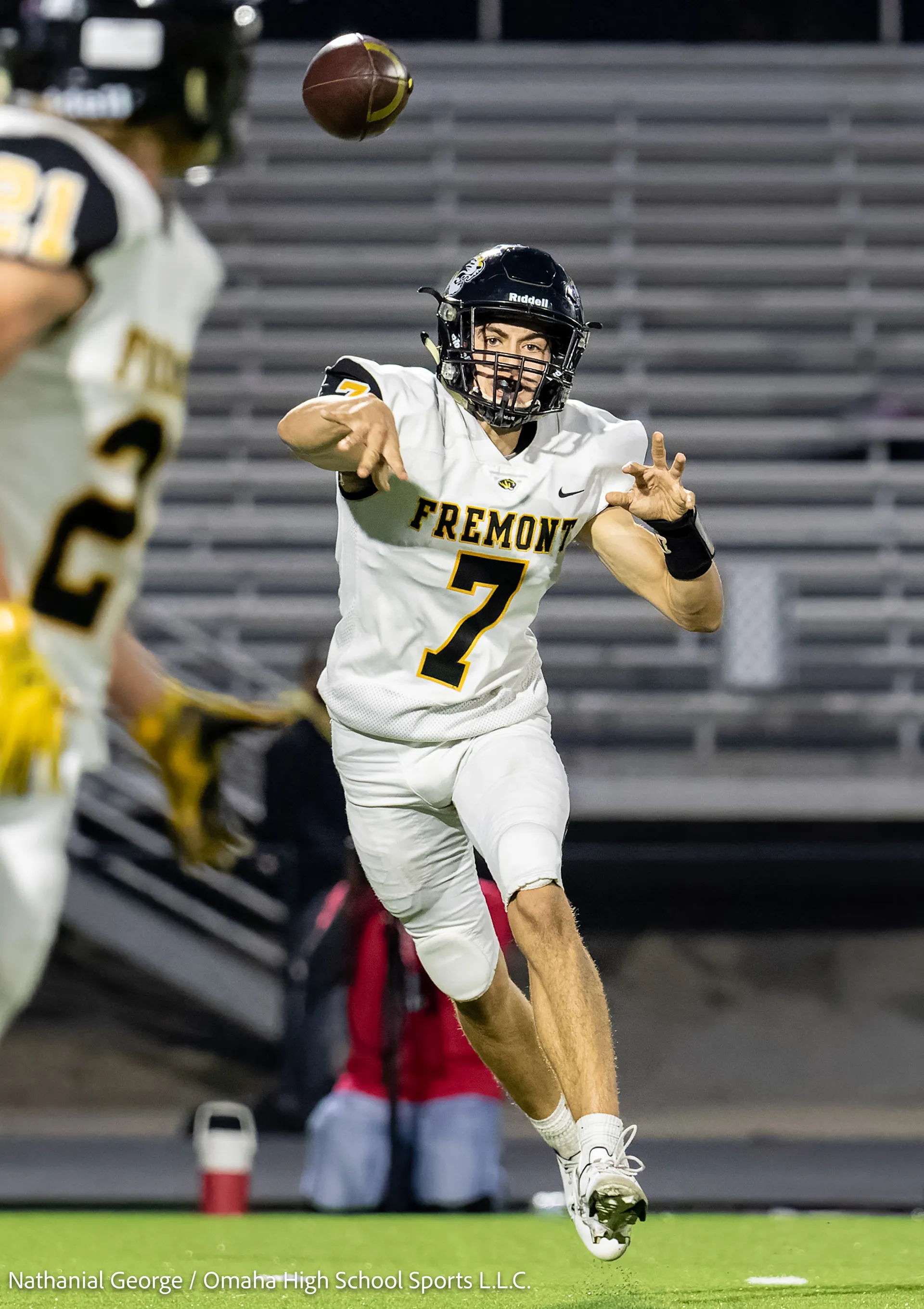 A Fremont quarterback in white, number 7, lets a pass go toward a teammate blurred in the foreground, with empty bleachers behind him.