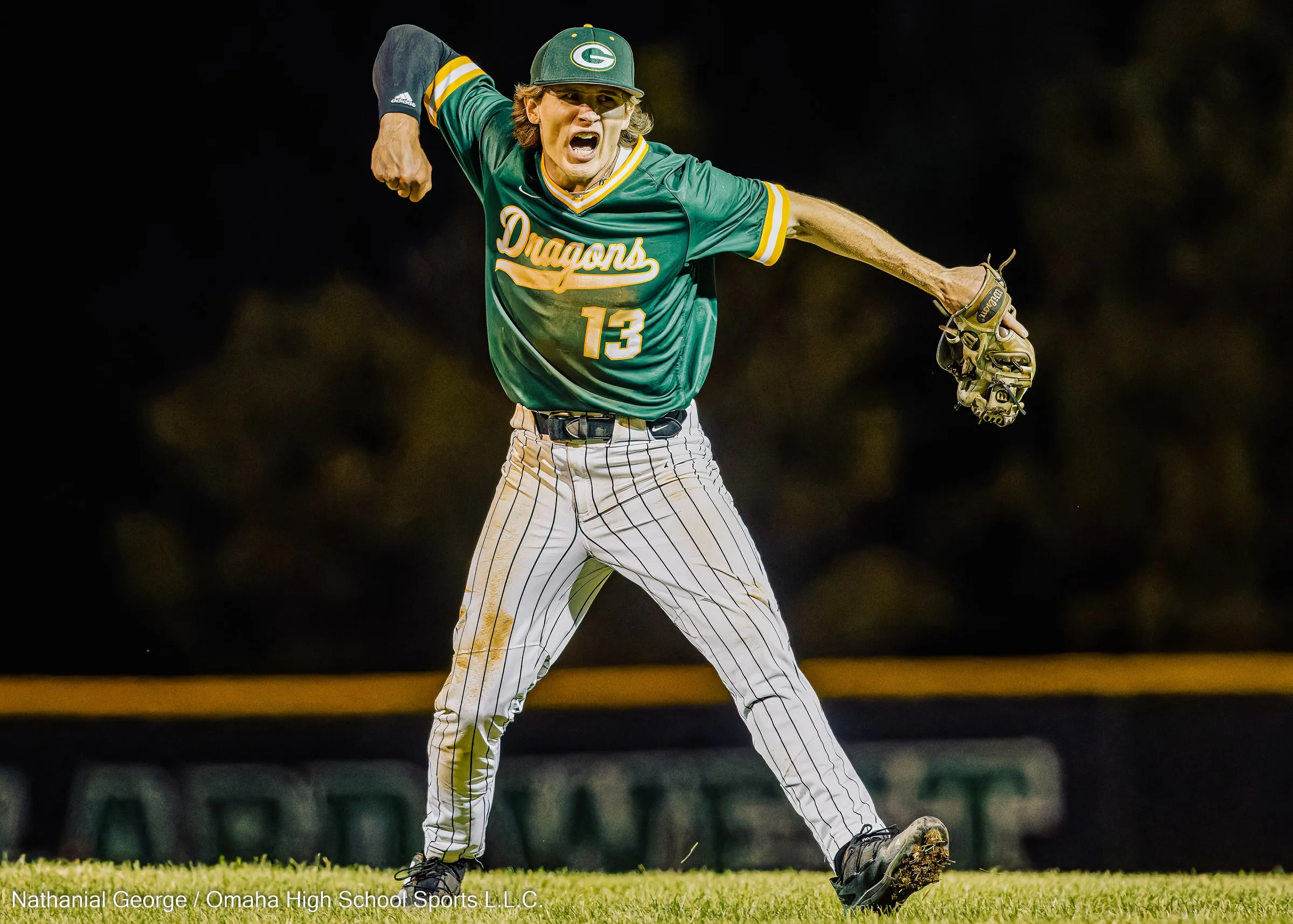 A Dragons infielder in green, number 13, shouts and pumps his fist with his glove flung wide, dirt on the knees of his pinstriped pants, at a night game.